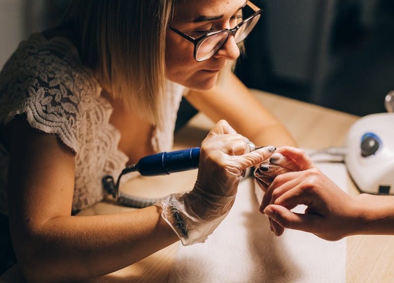 High angle of concentrated female master in glasses and gloves manicuring hand with drill of faceless client